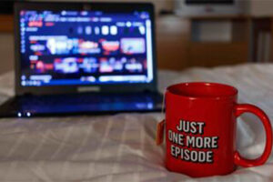 Open laptop with streaming service on screen resting on bed with mug in front of it with "Just One More Episode" message written on it