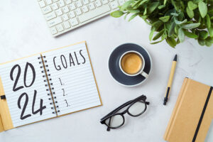 Desk surface with 2024 goals written in notebook, a coffee cup, glasses, a pen, a plant, and a keyboard in background