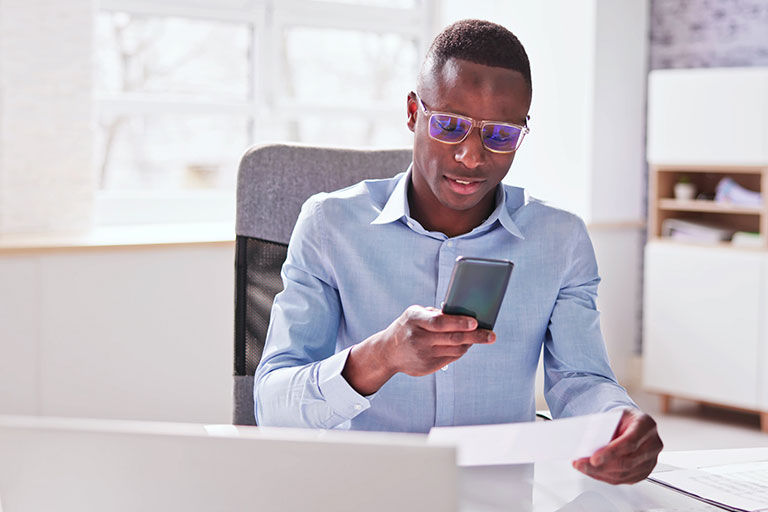Man of color sitting in office taking a picture of check with cell phone